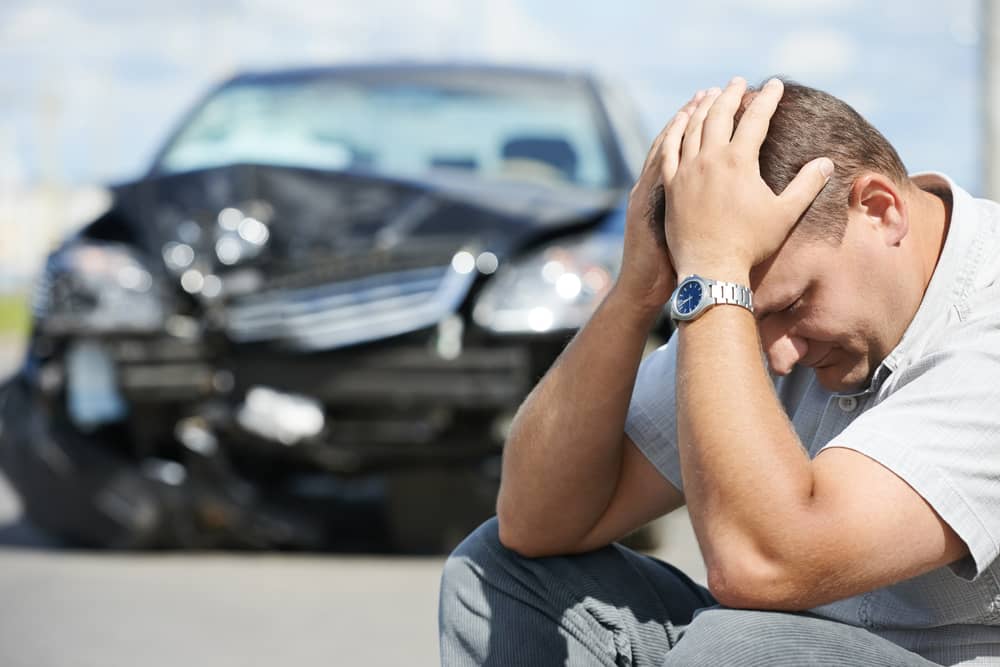 Auto accident victim sitting by road with a view of his wrecked car.