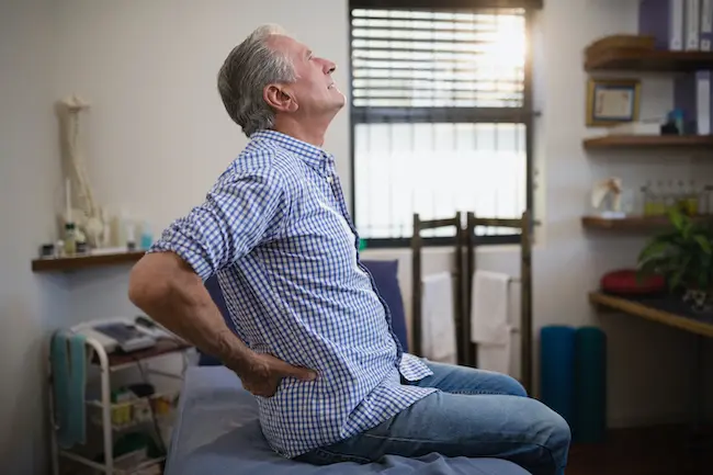 An elderly man sitting in a hospital ward suffering from lower back or hip pain.