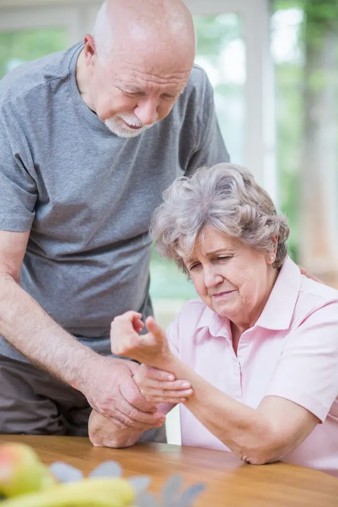 Senior woman massaging her hand in pain with husband trying to help.